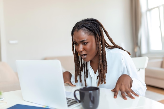 Verwirrte afrikanische Geschäftsfrau, die sich über Online-Problem-Spam-E-Mails oder gefälschte Internetnachrichten ärgert, die auf Laptop-Büroangestellte blicken und sich schockiert über festgefahrene Computer fühlen, die von Betrugsnachrichten oder Viren verwirrt sind