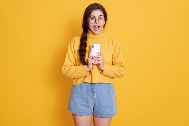 Verwirrte emotionale dunkelhaarige Frau hält Smartphone, öffnet den Mund weit, erstaunt über etwas Beeindruckendes, posiert isoliert auf gelber Wand, trägt Pullover und kurze Jeans.