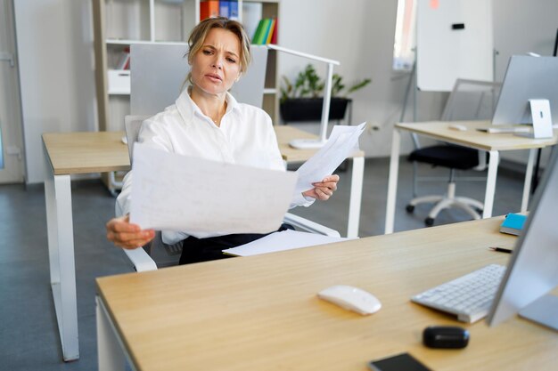 Verwirrte Geschäftsfrau, die Dokumente am Schreibtisch prüft