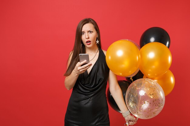 Verwirrte junge Frau im kleinen schwarzen Kleid mit Luftballons, mit Handy beim Feiern auf rotem Hintergrund. Valentinstag, guten Rutsch ins Neue Jahr, Geburtstagsmodell-Feiertagspartykonzept.