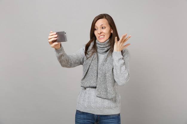 Verwirrte junge Frau im Pullover, Schal, die Hände ausbreitet, die Selfie-Aufnahmen auf dem Handy machen, die Videoanrufe einzeln auf grauem Hintergrund tätigen. Gesunde Mode Lifestyle Menschen Emotionen Konzept der kalten Jahreszeit.