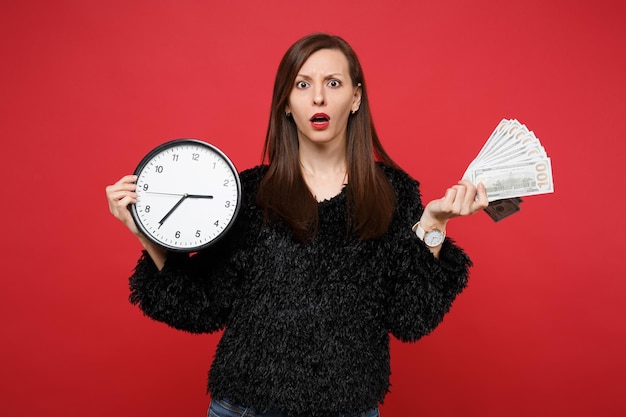 Verwirrte junge Frau in schwarzem Pelzpullover mit runder Uhr Fan von Geld in Dollar-Banknoten Bargeld isoliert auf rotem Hintergrund. Menschen aufrichtige Emotionen, Lifestyle-Konzept. Die Zeit wird knapp.