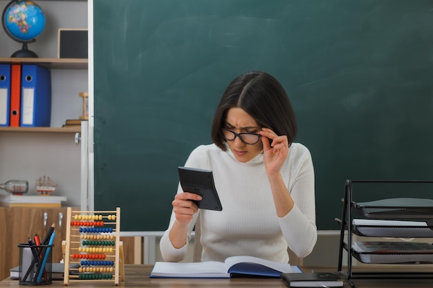 verwirrte junge lehrerin mit brille, die einen rechner hält und betrachtet, der am schreibtisch mit schulwerkzeugen im klassenzimmer sitzt