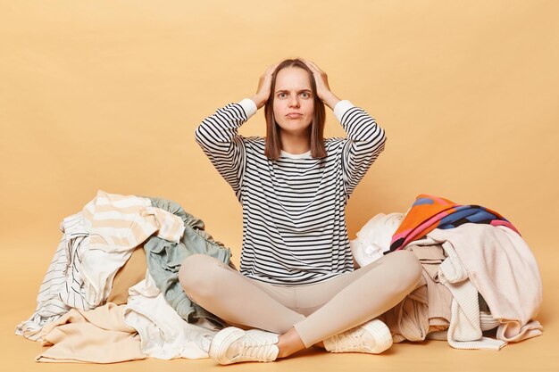 Foto verwirrte, schockierte, verwirrte kaukasische frau posiert in der nähe eines haufs mehrfarbiger, ungesortierter kleidung, isoliert über beige hintergrund, hält die hände auf dem kopf, muss viele kleidung sortieren.