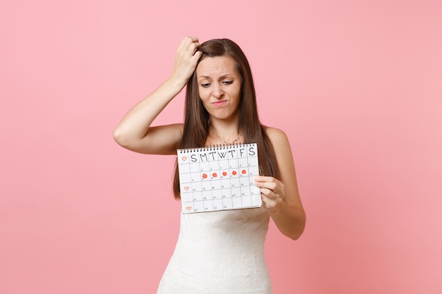 Verwirrte schuldige Frau im weißen Kleid, die sich im Kopf festklammert und auf den Kalender der weiblichen Perioden schaut, um die Menstruationstage zu überprüfen