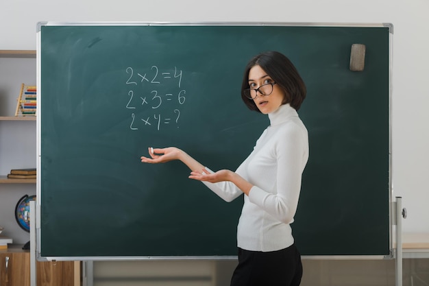 Verwirrte sich ausbreitende Hände Junge Lehrerin mit Brille steht vor der Tafel und schreibt im Klassenzimmer