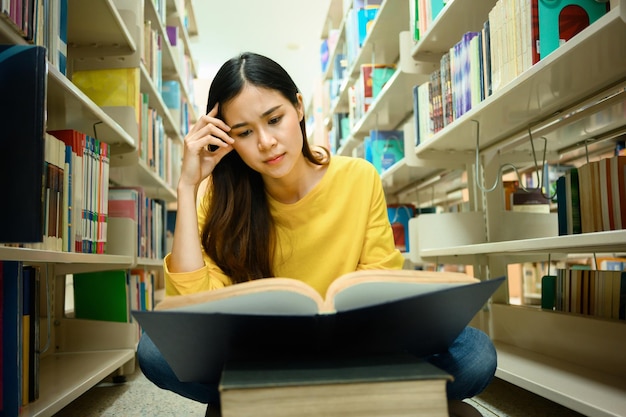 Verwirrte Studentin sitzt zwischen Bücherregalen in der Bibliothek und liest Bücher zum Lernen und Forschen