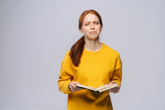 Verwirrte unzufriedene junge Studentin, die Buch hält und auf grau isoliertem Hintergrund weint Hübsches rothaariges Damenmodell mit gelbem Pullover, das emotional Gesichtsausdrücke zeigt