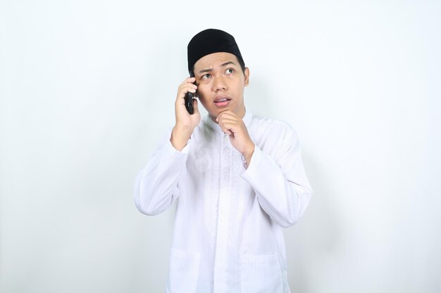Verwirrter asiatischer muslimischer Mann spricht am Telefon mit der Hand auf dem Kinn, isoliert auf weißem Hintergrund