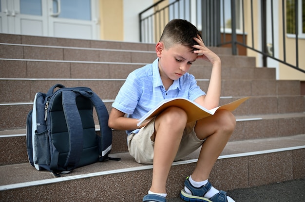 Verwirrter Grundschüler, Schüler, Schuljunge, der seine Hausaufgaben auf einem Notebook macht, während er auf der Treppe einer Schuleinrichtung sitzt.