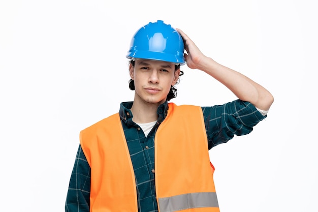 Verwirrter junger männlicher Ingenieur mit Schutzhelm und Warnweste, der in die Kamera blickt und die Hand auf dem Kopf isoliert auf weißem Hintergrund hält