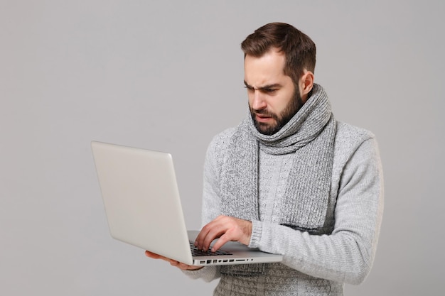 Verwirrter junger Mann im grauen Pullover, Schal isoliert auf grauem Hintergrund. Gesunder Lebensstil kranke Krankheit, Online-Behandlungsberatung, Konzept der kalten Jahreszeit. Mock-up-Kopienbereich. Halten Sie einen Laptop-PC-Computer.