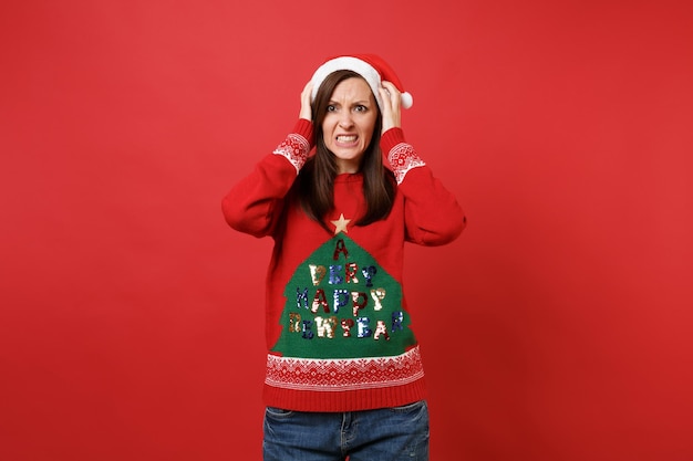 Verwirrtes, beschäftigtes junges Santa-Mädchen in Strickpullover, Weihnachtsmütze, die Hände auf den Kopf legt, isoliert auf rotem Wandhintergrund. Frohes neues Jahr 2019 Feier Urlaub Party Konzept. Kopieren Sie Platz.