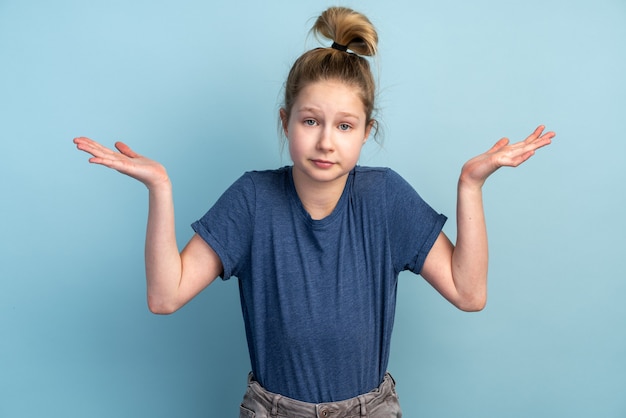 Verwirrtes junges Mädchen an einer blauen Wand breitet seine Arme aus