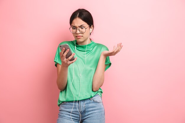 Verwirrtes junges Mädchen mit runder Brille mit Kopfhörern, während es das Smartphone isoliert über rosafarbener Wand hält?