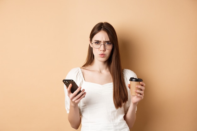 Verwirrtes Mädchen mit Brille runzelt die Stirn, nachdem es das Handy mit der Kaffeetasse im Stehen gelesen hat.