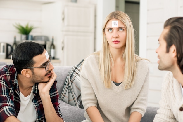 Verwirrtes Mädchen mit langen blonden Haaren, die versuchen, ihren Namen auf Aufkleber zu erraten, während sie Spiel mit ihren Freunden in der Freizeit spielen