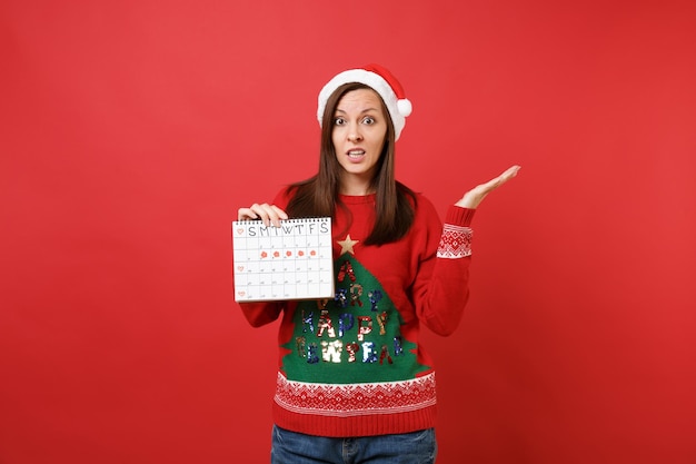 Verwirrtes Santa-Mädchen, das den Kalender der weiblichen Perioden hält, um die Menstruationstage einzeln auf rotem Hintergrund zu überprüfen. Medizinisches Gesundheitswesen, gynäkologisch. Frohes neues Jahr 2019 Feier Urlaub Party Konzept.