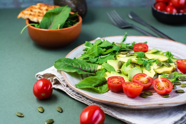 Verzehrfertiger Salat mit Avocado, Tomaten, Rucola, Mizuna, Mangold und Kürbiskernen auf einem Teller auf einem grünen Tisch. Vegetarische Vitaminnahrung. Platz kopieren