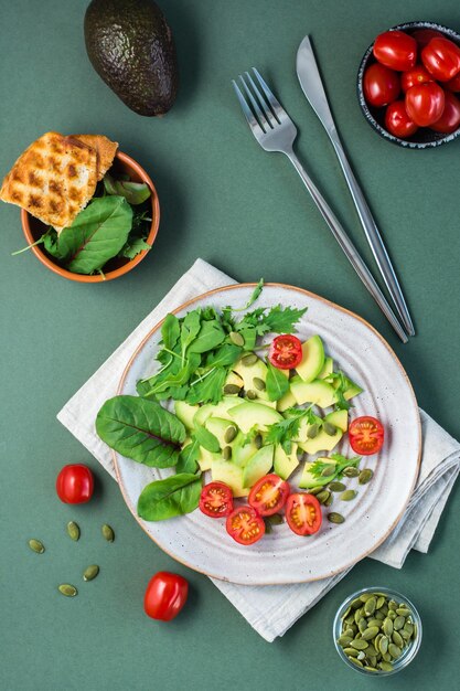 Verzehrfertiger Salat mit Avocado, Tomaten, Rucola, Mizuna, Mangold und Kürbiskernen auf einem Teller und Besteck auf einem grünen Tisch. Vegetarische Vitaminnahrung. Ansicht von oben und vertikal