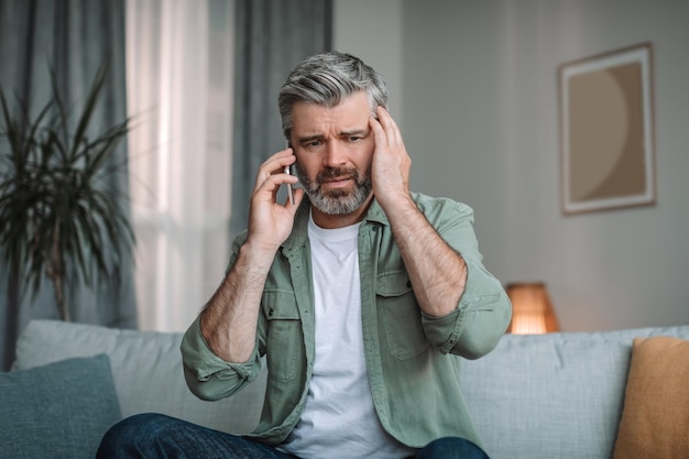 Verzweifelter trauriger älterer europäischer Mann mit Bart spricht per Smartphone und bekam schlechte Nachrichten im Innenraum