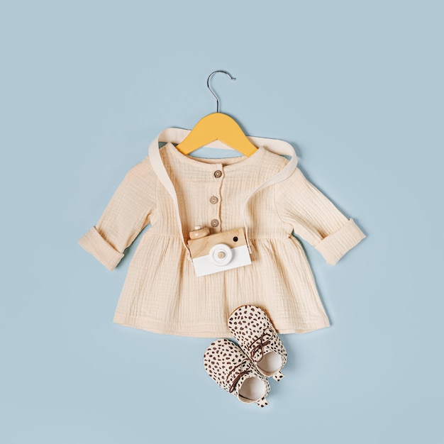 Vestido branco em cabide com câmera de brinquedo e sapatos. Conjunto de roupas de bebê e acessórios para a primavera, outono ou verão em fundo azul claro. Roupa de moda infantil. Camada plana, vista superior