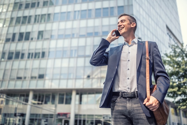 Vestido de negócios casual para homem que fala ao telefone celular em frente ao prédio de escritórios sorrindo.