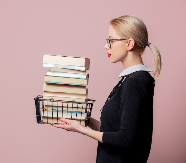 Vestido e óculos pretos de mulher loira segurando uma cesta de compras com livros na parede rosa