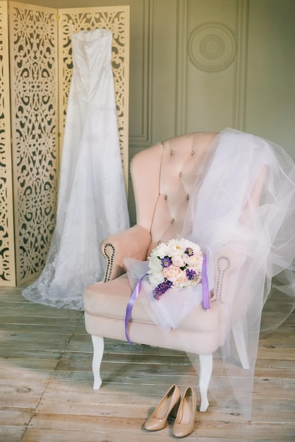 Vestido de novia de bellas artes con encaje en gabinete de madera pantalla plegable ramo de peonías en silla rosa cubierta con tonos pastel de tela suave Zapatos en el piso en la preparación matutina de la novia del hotel