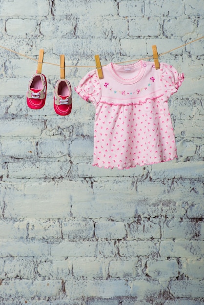 Vestido rosa bebé y zapatos rojos para la niña se secan en una cuerda contra una pared de ladrillo blanco.