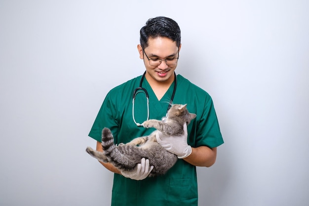 Veterinario asiático sonriente con batas verdes y estetoscopio sosteniendo gato