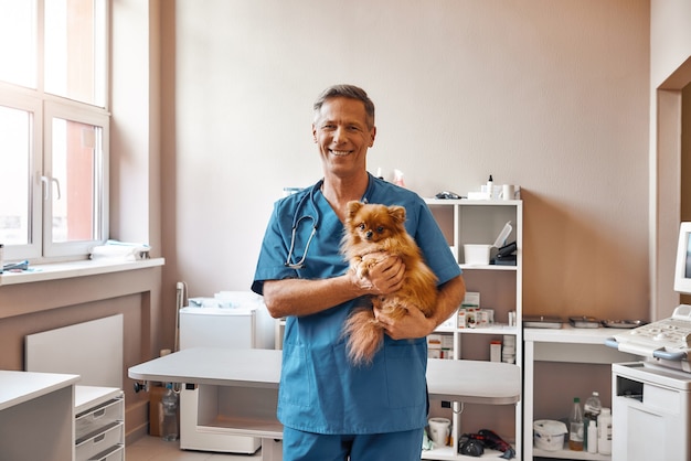 El veterinario en uniforme de trabajo sostiene un perrito y sonríe a la cámara mientras está de pie en la clínica veterinaria