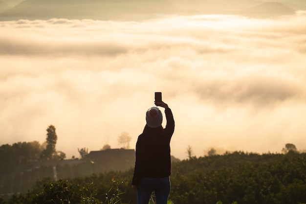 Viagem de inverno relaxa conceito de férias. jovem viajante asiática com blusa e chapéu de lã tirando foto e selfie com telefone celular na montanha com névoa ao nascer do sol em mae hong son, tailândia