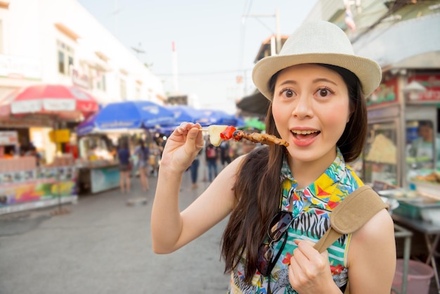 viagem de jovem empresária asiática trabalhando em Bangkok alegre comendo kebabs de churrasco de carne de frango local no famoso mercado de rua ao ar livre nas férias de Tailândia.