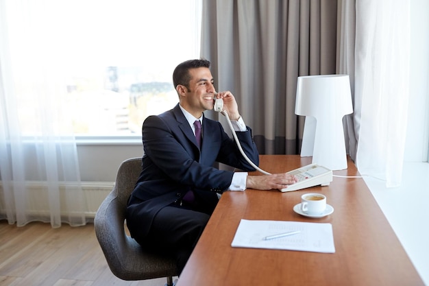 viagem de negócios, pessoas e conceito de comunicação - feliz sorridente empresário com café e papéis chamando no telefone de mesa no quarto do hotel