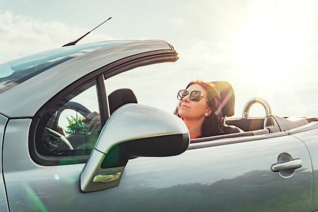 Viagem de verão ela viaja para um show com teto aberto mulher satisfeita sentada relaxada no carro contra o céu com sol brilhante conceito de viajar de carro férias de verão prazer de dirigir