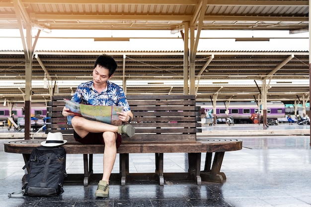 Viagem ou viagem de estilo de vida de conceito jovem viajante asiático sentado em um banco de madeira e olhando para o mapa enquanto espera o trem chegar à estação