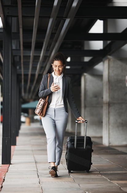 Viagem telefônica e mala com uma mulher de negócios andando em um estacionamento de aeroporto ao ar livre na cidade Bagagem móvel e comutação com uma jovem funcionária em uma viagem internacional a trabalho