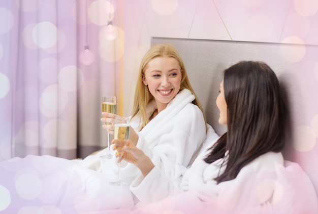 viagens, amizade, despedida de solteira e conceito de pessoas - namoradas sorridentes em roupões de banho com taças de champanhe na cama sobre fundo de luzes de férias