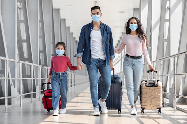 Viagens durante a pandemia. família árabe de três usando máscaras médicas protetoras andando com malas no terminal do aeroporto, pais árabes com filha pequena de mãos dadas enquanto vai para o portão