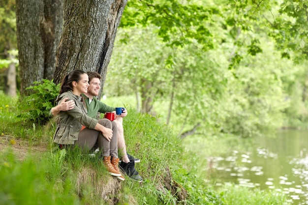 viagens, turismo, caminhada, camping e conceito de pessoas - casal feliz com xícaras bebendo chá e abraçando na natureza na margem do rio