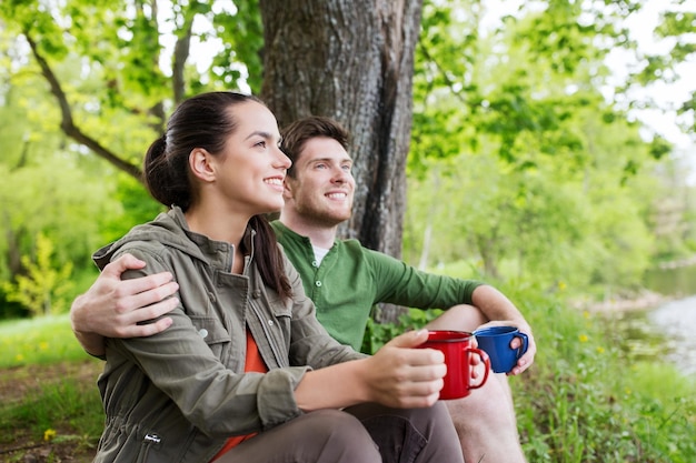 viagens, turismo, caminhada, camping e conceito de pessoas - casal feliz com xícaras bebendo chá na natureza na margem do rio