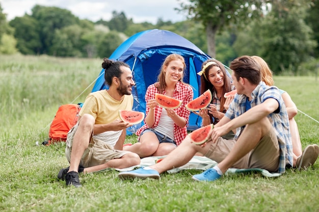 viagens, turismo, caminhada, piquenique e conceito de pessoas - grupo de amigos felizes com tenda comendo melancia no acampamento