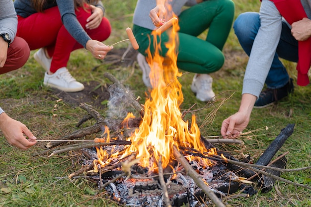 Viagens, turismo, caminhada, piquenique e pessoas - grupo de amigos felizes fritando salsichas na fogueira