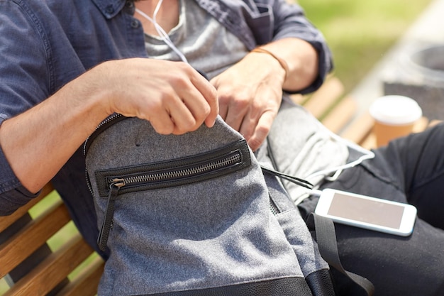 viagens, turismo, estilo de vida e conceito de pessoas - homem com fones de ouvido e óculos de sol sentado no banco da cidade e procurando algo em sua mochila