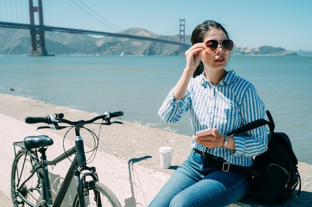 viajante asiática está olhando para a distância enquanto usa o aplicativo gps no telefone durante uma pausa do ciclismo à beira-mar perto da ponte golden gate na Califórnia