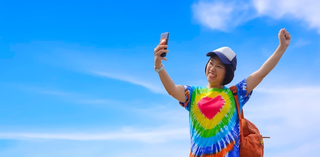 Viajante asiática usando smartphone tirando selfie com levantar a mão contra o fundo do céu azul