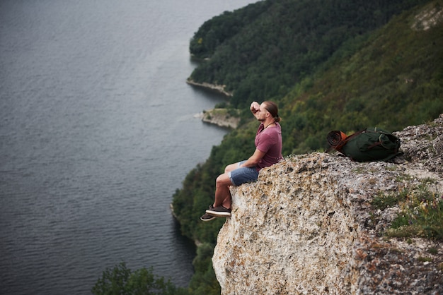 Viajante com mochila, sentado no topo da montanha, apreciando a vista acima da superfície da água. liberdade e conceito de estilo de vida ativo