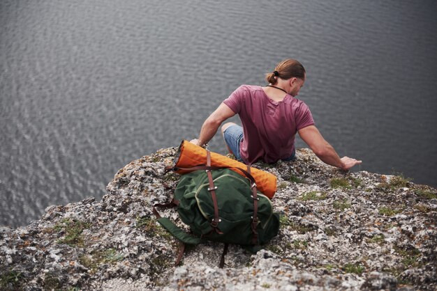 Viajante com mochila, sentado no topo da montanha, apreciando a vista acima da superfície da água. liberdade e conceito de estilo de vida ativo