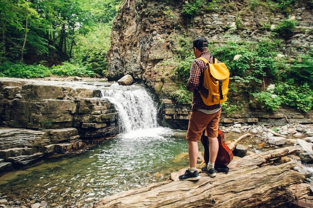 Viajante com uma mochila e um violão em pé no tronco de uma árvore contra uma cachoeira